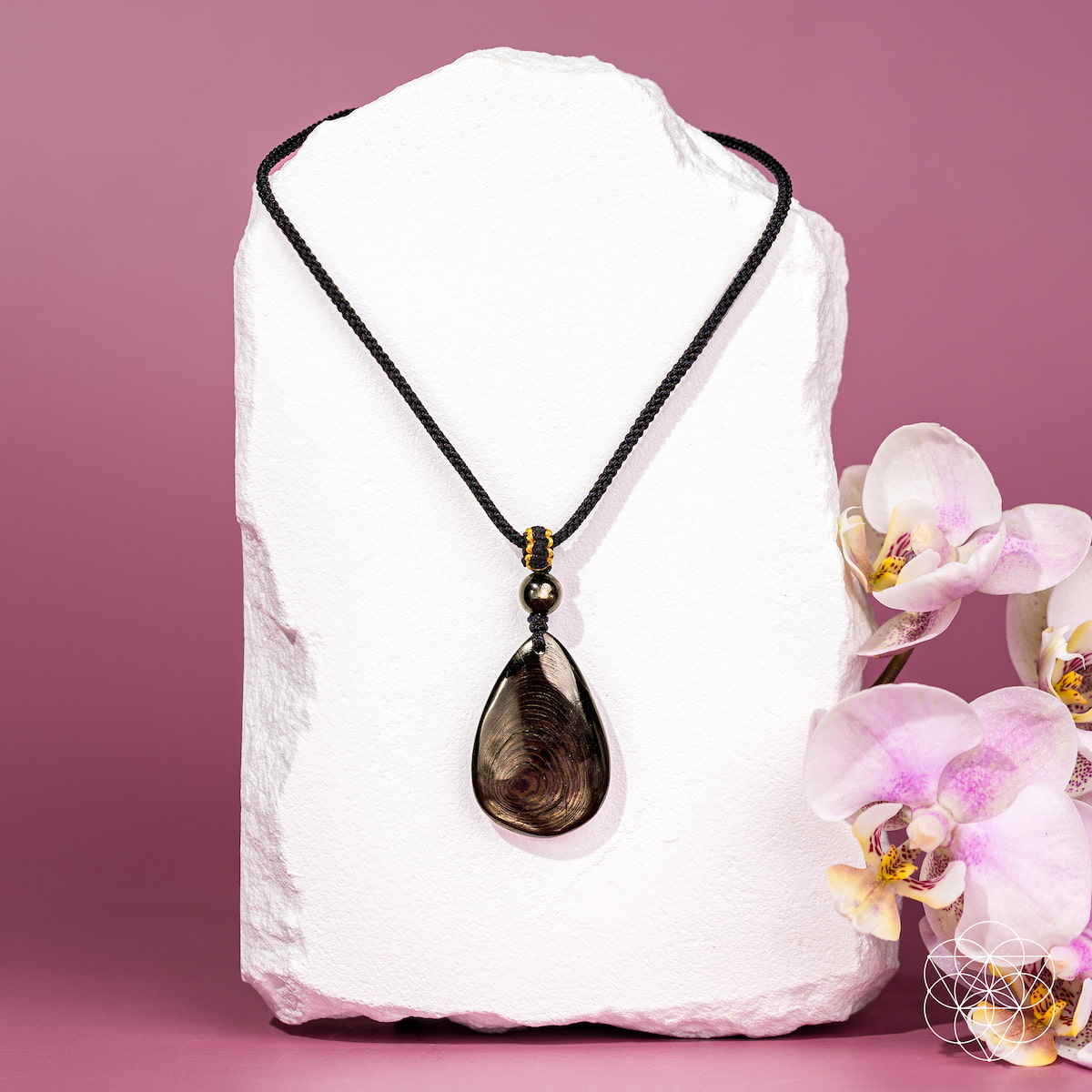 The Hypersthene Millionaire Manifestation Pendant by Conscious Items features a dark, swirled design on a black cord, presented on a white stone stand with orchids—a striking piece for wealth manifestation against a pink backdrop.