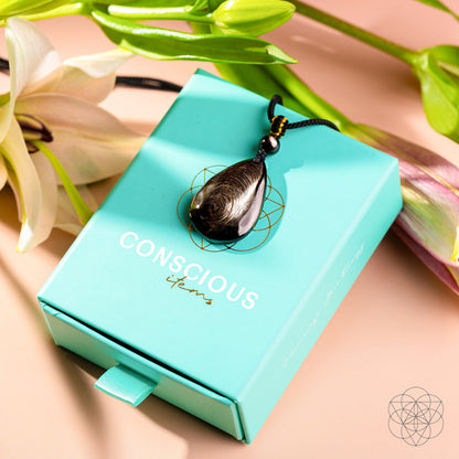 The Hypersthene Millionaire Manifestation Pendant by Conscious Items, a teardrop-shaped stone on a black cord, rests atop the brand’s turquoise box amid white and pink lilies—an elegant symbol of wealth manifestation.