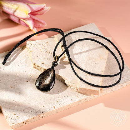 The Hypersthene Millionaire Manifestation Pendant by Conscious Items, a wealth-attracting black gemstone, is displayed on beige stone slabs with a pink lily in the background against a soft pink surface.