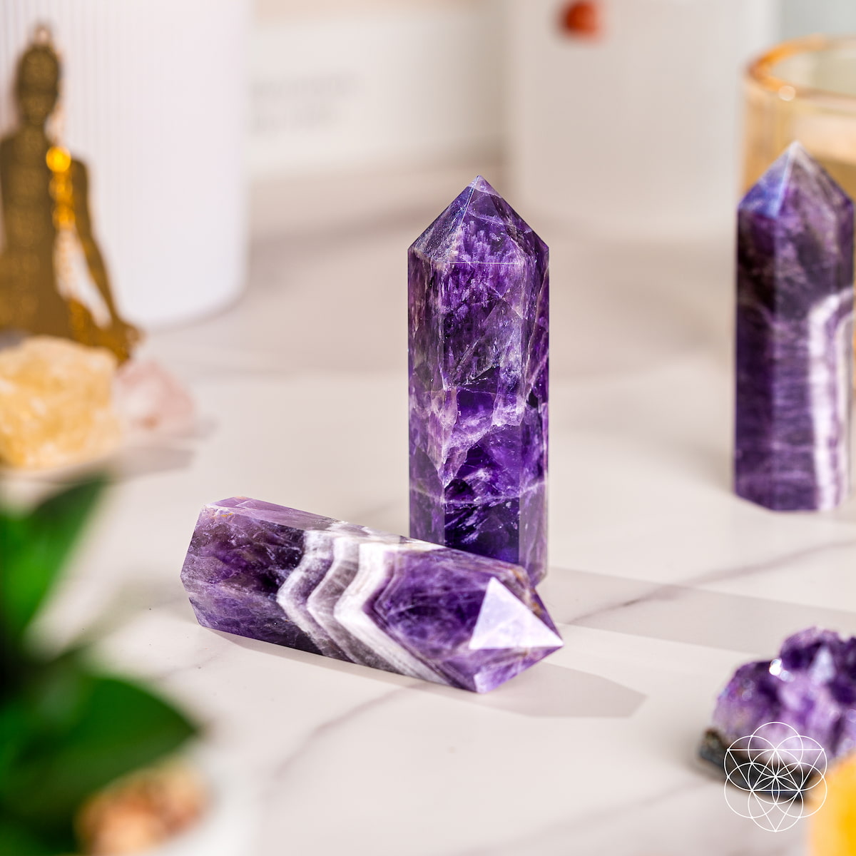 Three polished Cycle Breaker - Chevron Amethyst Points of Transformation from Conscious Items, known for stress relief and sleep support, are arranged on a white surface with a blurred green plant, gold figurine, and candles in the background.
