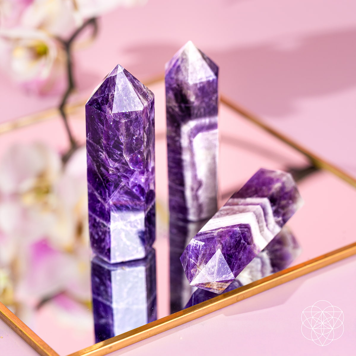 Three Cycle Breaker - Chevron Amethyst Points of Transformation from Conscious Items sit on a gold-edged mirrored tray, reflecting purple and white hues. A blurred orchid and logo in the background enhance this calming stone for sleep.