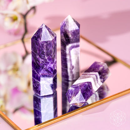 Three Cycle Breaker - Chevron Amethyst Points of Transformation from Conscious Items sit on a gold-edged mirrored tray, reflecting purple and white hues. A blurred orchid and logo in the background enhance this calming stone for sleep.