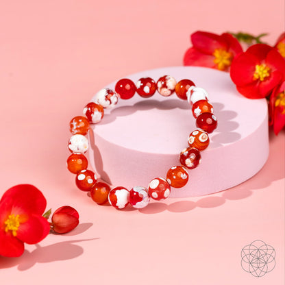 The Feminine Force – Carnelian Orca Agate Bracelet by Conscious Items, with red and white marbled beads, is showcased on a round pink platform amid vibrant red flowers against a pastel pink background.