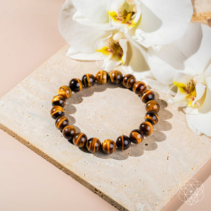 The Built to Win – 7A Grade Tiger’s Eye Bracelet by Conscious Items is showcased on a square beige stone slab, complemented by two white orchid flowers in the background.