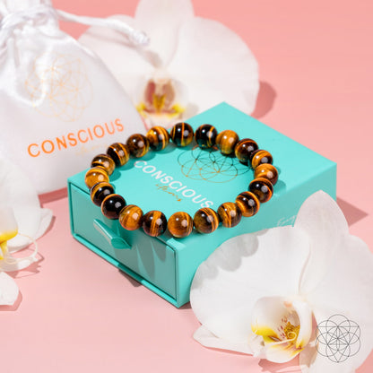 The Built to Win – 7A Grade Tiger’s Eye Bracelet by Conscious Items rests on a turquoise Conscious Items box, surrounded by white orchids and a white Conscious branded drawstring pouch, all displayed on a pink surface.