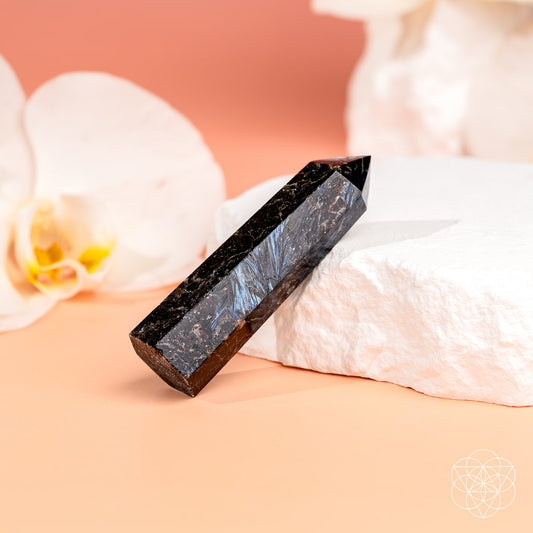 The "This Is Your Breakthrough – Rare Arfvedsonite Point" from Conscious Items, celebrated for its breakthrough energy, is displayed on a white stone slab with white orchids and a soft peach background.