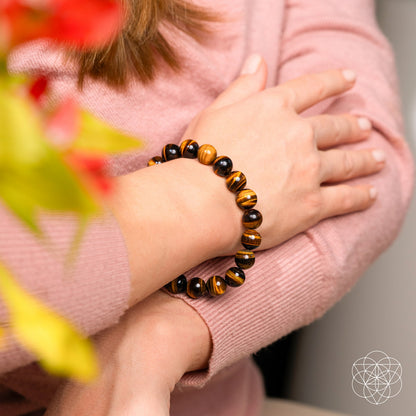 A person in a pink sweater crosses their arms, wearing the Conscious Items Built to Win – 7A Grade Tiger’s Eye Bracelet with brown and black striped stones. Red and green blurred flowers are in the foreground; a white geometric logo sits bottom right.