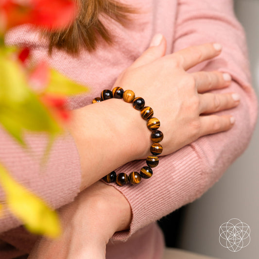 A person in a pink sweater crosses their arms, wearing the Conscious Items Built to Win – 7A Grade Tiger’s Eye Bracelet with brown and black striped stones. Red and green blurred flowers are in the foreground; a white geometric logo sits bottom right.