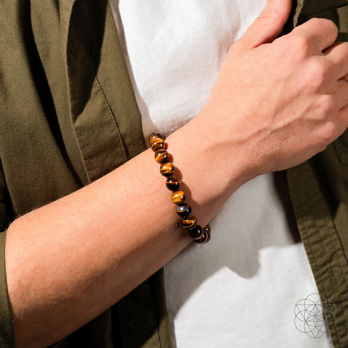 A person in a white shirt and olive green jacket wears the Conscious Items “Built to Win” 7A Grade Tiger’s Eye Bracelet, featuring polished brown and gold stones, on their left wrist.