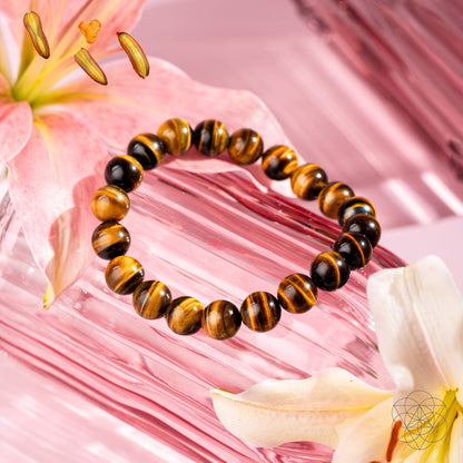 The Built to Win – 7A Grade Tiger’s Eye Bracelet by Conscious Items is displayed on a pink textured surface, surrounded by pale pink and white lilies with floating stamens.