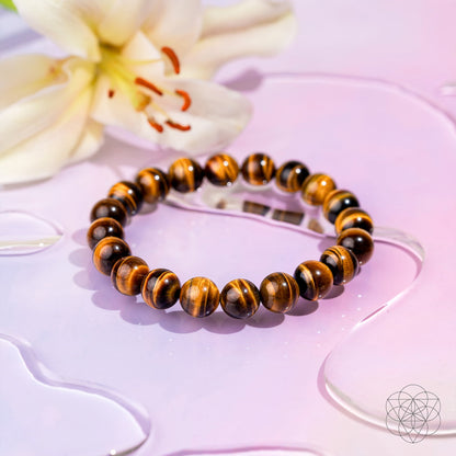 The Built to Win – 7A Grade Tiger’s Eye Bracelet by Conscious Items rests on a glossy pink surface with water droplets, beside a pale yellow lily flower.