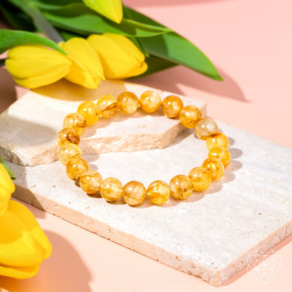 The Master Reset – Golden Healer Quartz Bracelet by Conscious Items is showcased on a light stone slab with vibrant yellow tulips, set against a soft pink backdrop.