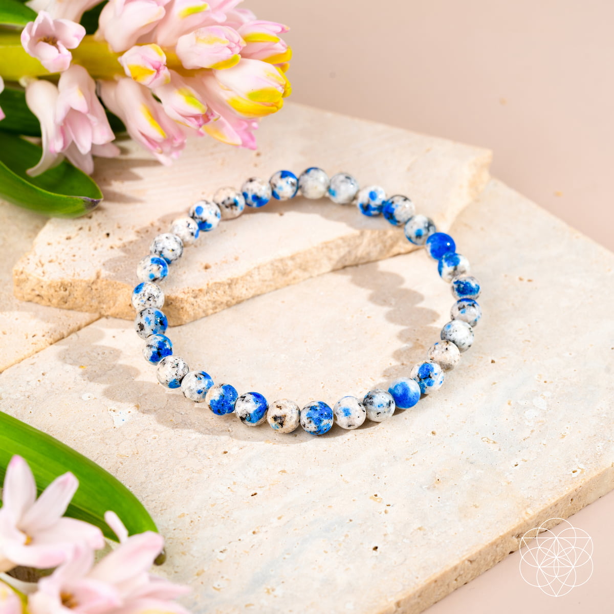 The Rare K2 Stone Bracelet of Spiritual Adventure | 6mm by Conscious Items is shown on light stone slabs with pink flowers and green leaves, highlighting its blue and white marbled beads in soft natural light.