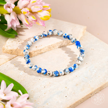 The Rare K2 Stone Bracelet of Spiritual Adventure | 6mm by Conscious Items is shown on light stone slabs with pink flowers and green leaves, highlighting its blue and white marbled beads in soft natural light.