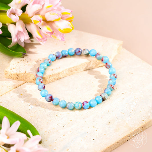 The Empath’s Shield – Aqua Terra Jasper Bracelet | 6mm by Conscious Items, featuring blue, pink, and purple beads, rests among hyacinth flowers and leaves—ideal for emotional detox and auric protection in a soft, bright setting.