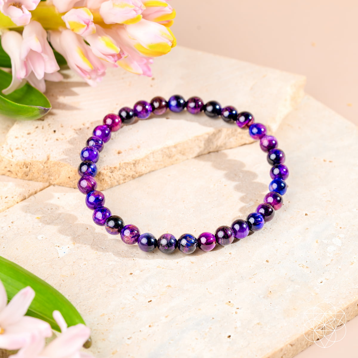 The Destiny Activator - All-Galaxy Tiger’s Eye Bracelet | 6mm by Conscious Items, featuring purple and blue gemstone beads, is shown on a light stone surface with pink and yellow flowers as decoration.