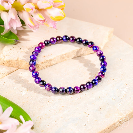 The Destiny Activator - All-Galaxy Tiger’s Eye Bracelet | 6mm by Conscious Items, featuring purple and blue gemstone beads, is shown on a light stone surface with pink and yellow flowers as decoration.
