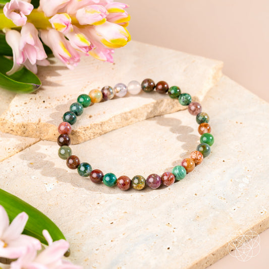 The Energy Bracelet | 6mm by Conscious Items, featuring multicolored round Indian Agate beads, is displayed on beige stone slabs with pink and yellow tulips in soft lighting, creating a calm and elegant atmosphere of emotional strength.