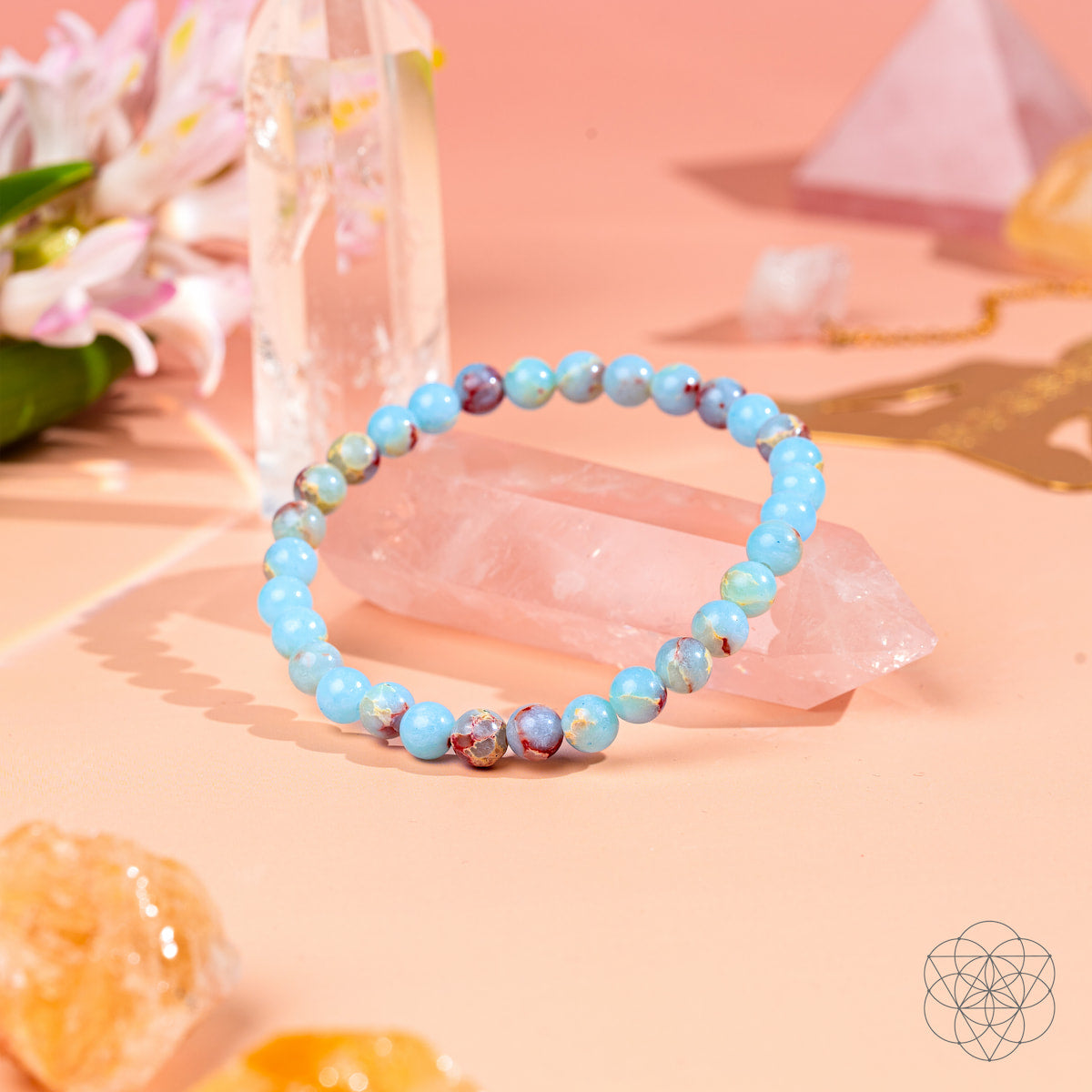 The Empath’s Shield – Aqua Terra Jasper Bracelet | 6mm by Conscious Items, featuring blue and marbled stones, rests on rose quartz with crystals, flowers, and geometric accents—promoting auric protection on a softly lit pink surface.