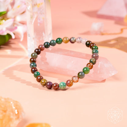 The Energy Bracelet | 6mm by Conscious Items, crafted from multicolored polished stones including Indian Agate for emotional strength, is displayed on clear quartz and surrounded by flowers and crystals on a soft pink surface.