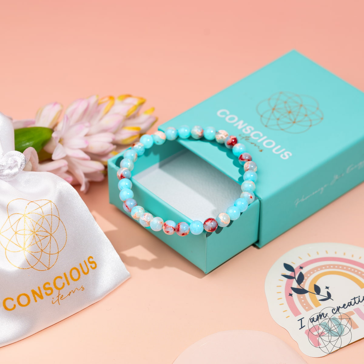The Empath’s Shield – Aqua Terra Jasper Bracelet | 6mm by Conscious Items rests on a light blue Conscious Items box, alongside a white satin pouch with gold logo, a flower, and decorative stickers on a peach-colored surface.