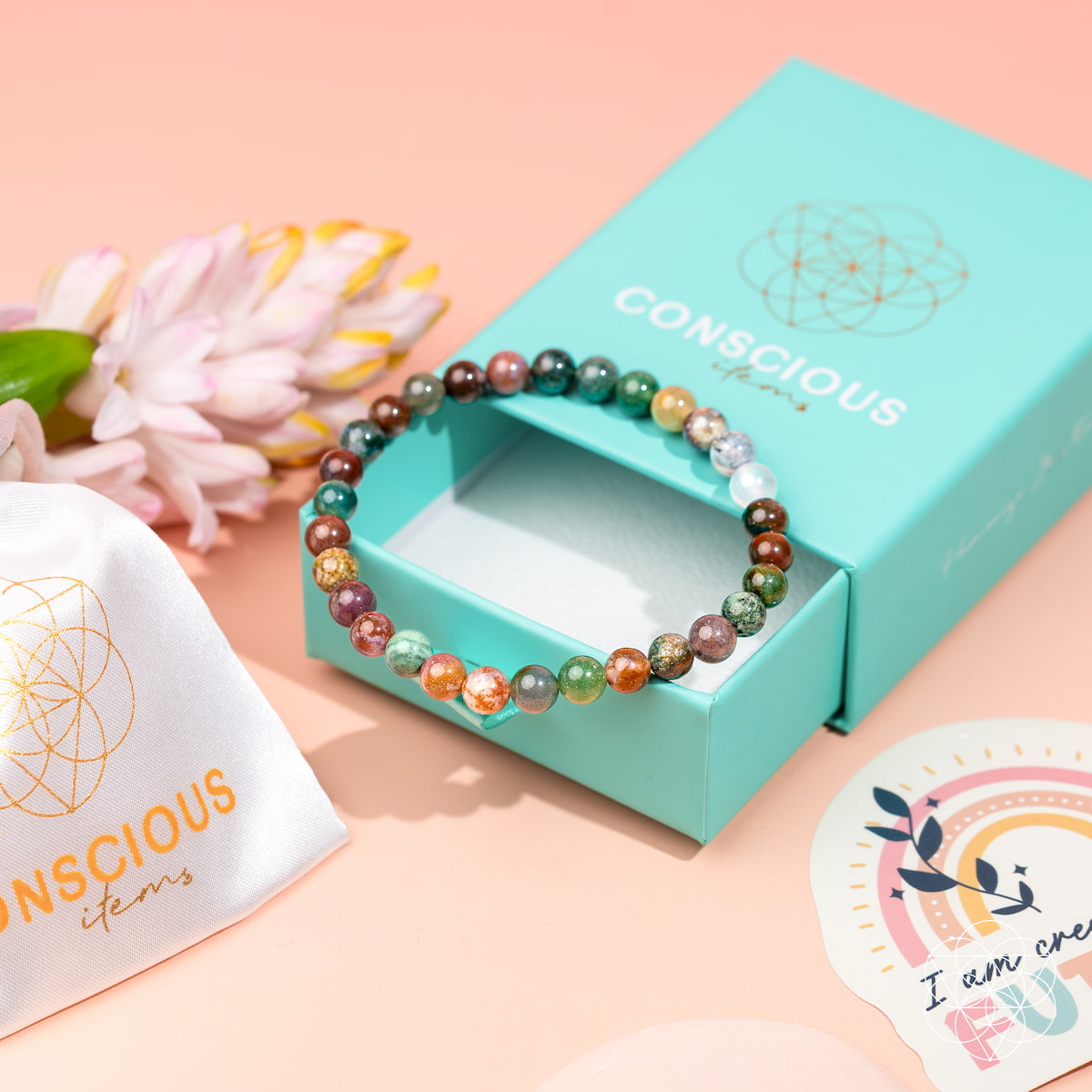 The Energy Bracelet | 6mm by Conscious Items, featuring multicolored Indian Agate beads, is displayed on a turquoise box with a white gold-lettered pouch, decorative sticker, and flower—perfect for an energy boost on a soft pink surface.