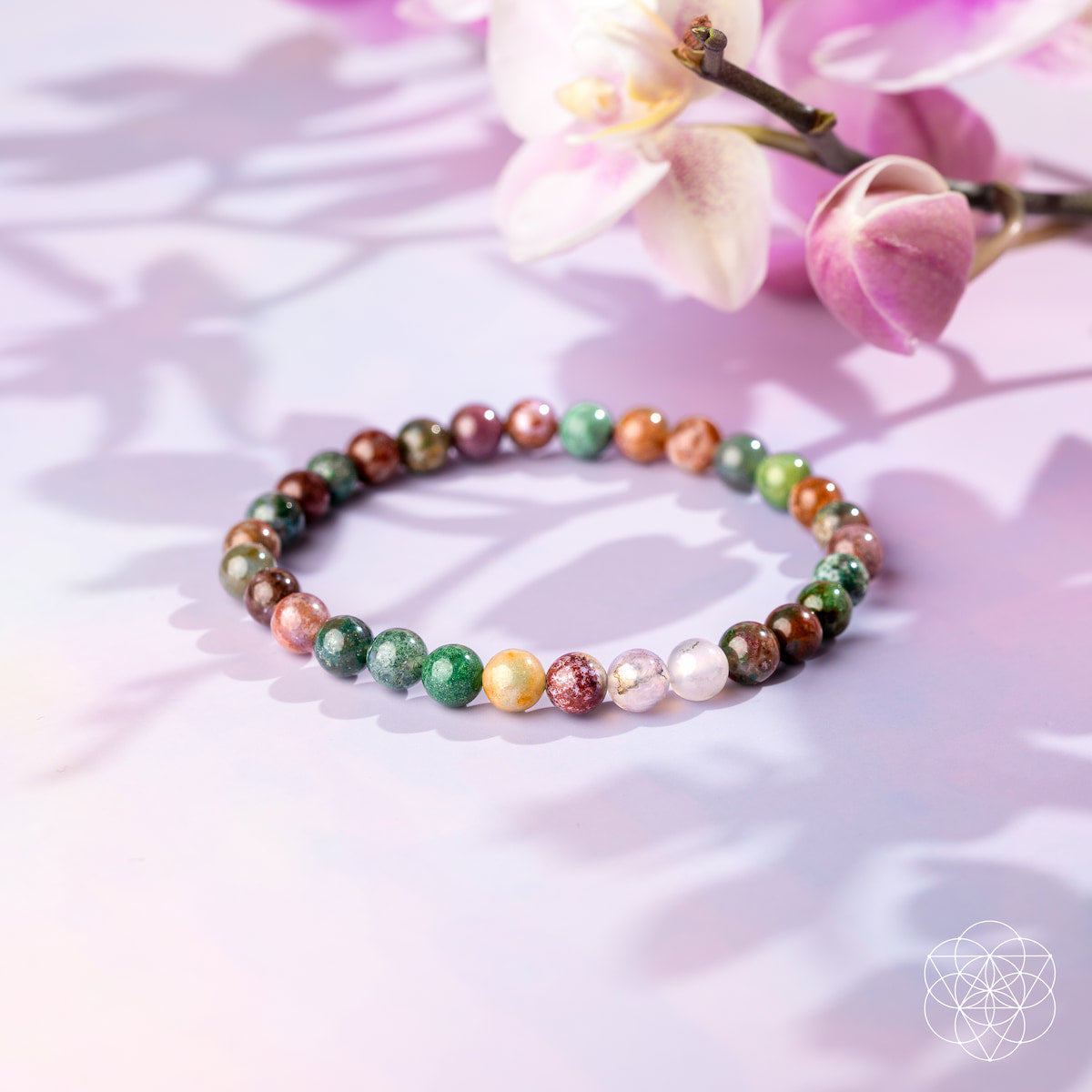 The Energy Bracelet | 6mm by Conscious Items, featuring multicolored Indian Agate beads for energy and emotional strength, is displayed on a soft pastel surface with pink and white orchids casting gentle shadows. A geometric white logo appears below.