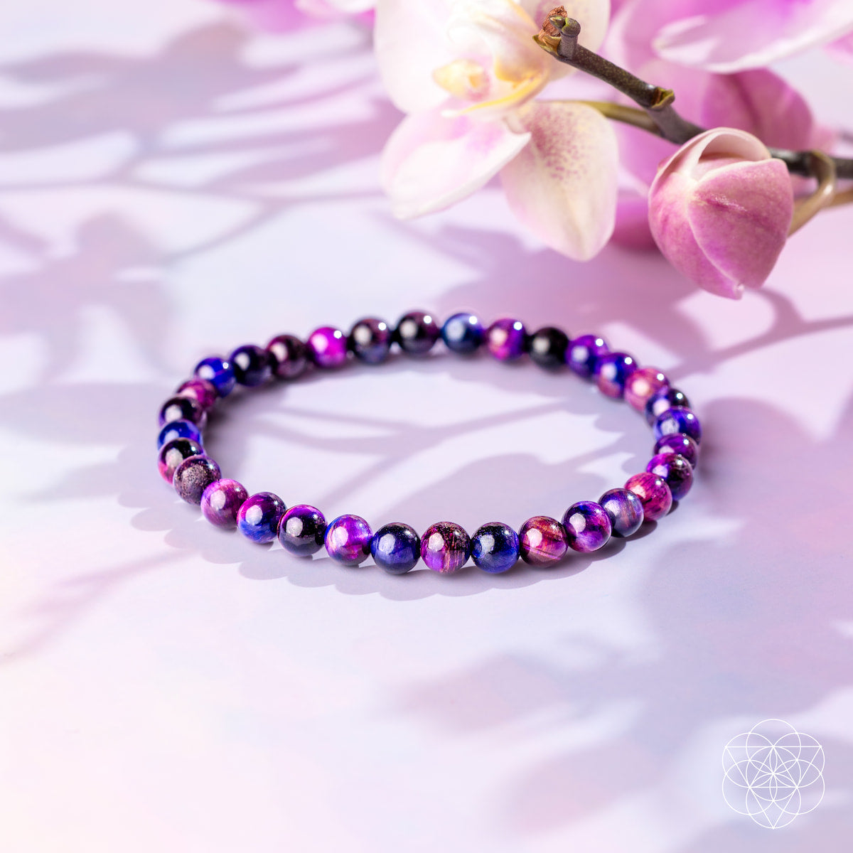 The Destiny Activator - All-Galaxy Tiger’s Eye Bracelet | 6mm by Conscious Items, featuring glossy purple, pink, and black beads, is displayed on a pastel surface with a pink orchid and gentle shadows in the background.