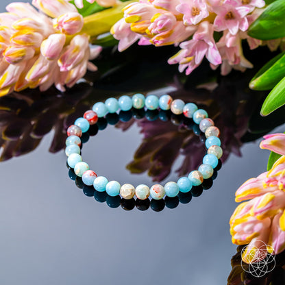 The Empath’s Shield – Aqua Terra Jasper Bracelet | 6mm by Conscious Items features light blue and speckled beads, displayed on a reflective black surface with pink flowers—ideal for emotional detox and subtle auric protection.