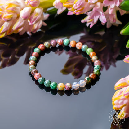 The Energy Bracelet | 6mm by Conscious Items, crafted from multicolored Indian Agate beads, rests on a glossy black surface with pink and yellow flowers. Its shimmering reflection appears below, alongside a white geometric logo in the corner.