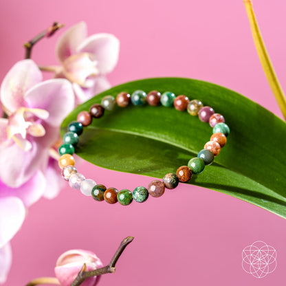 The Energy Bracelet | 6mm by Conscious Items, featuring vibrant Indian Agate beads, is displayed on a green leaf with pink orchids and a soft pink backdrop. A white geometric logo in the corner represents emotional strength and positivity.