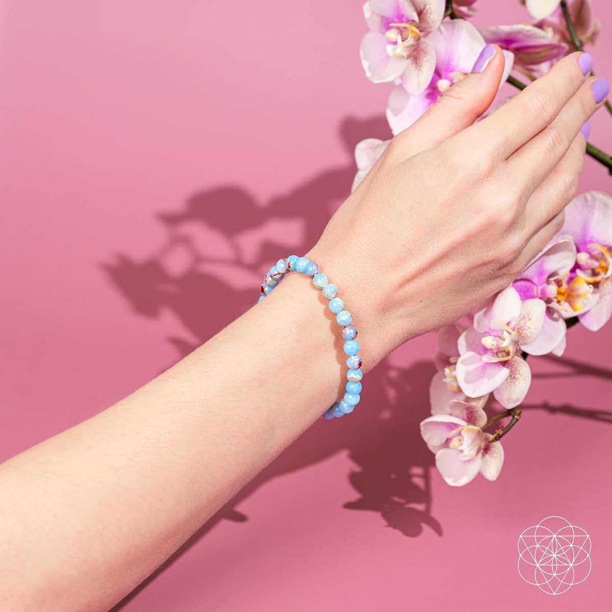 A hand wears the Empath’s Shield – Aqua Terra Jasper Bracelet | 6mm by Conscious Items, reaching toward pink and white orchids against a pink backdrop with a white geometric logo, evoking emotional detox and gentle auric protection.