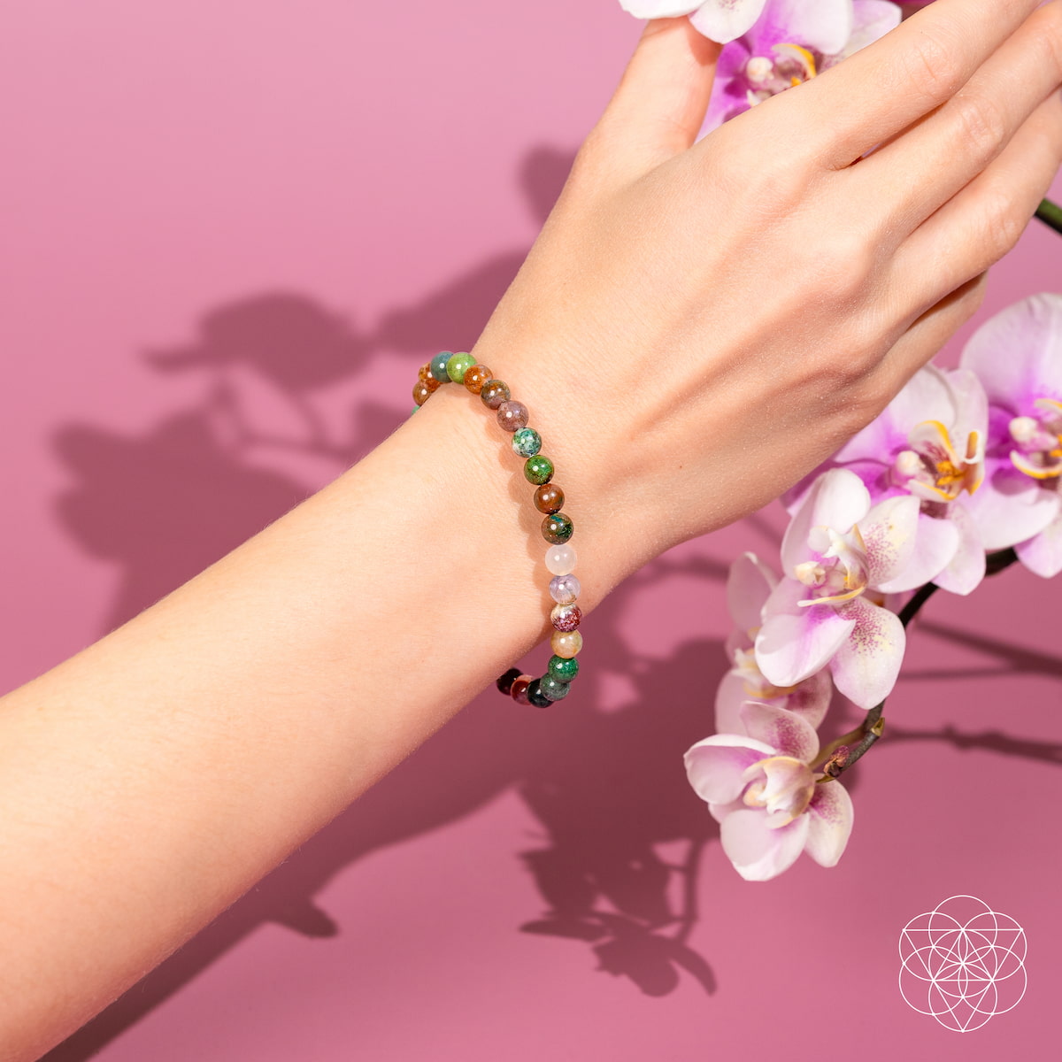 A hand adorned with The Energy Bracelet | 6mm by Conscious Items, featuring Indian Agate beads, is extended over a pink background near blooming white and purple orchids, symbolizing emotional strength.