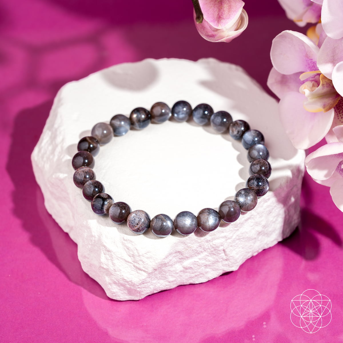 The Empath’s Cord Cutter – Black Moonstone Bracelet by Conscious Items rests on a white textured rock, set against a vibrant pink background with orchid flowers—perfect for symbolizing new beginnings.