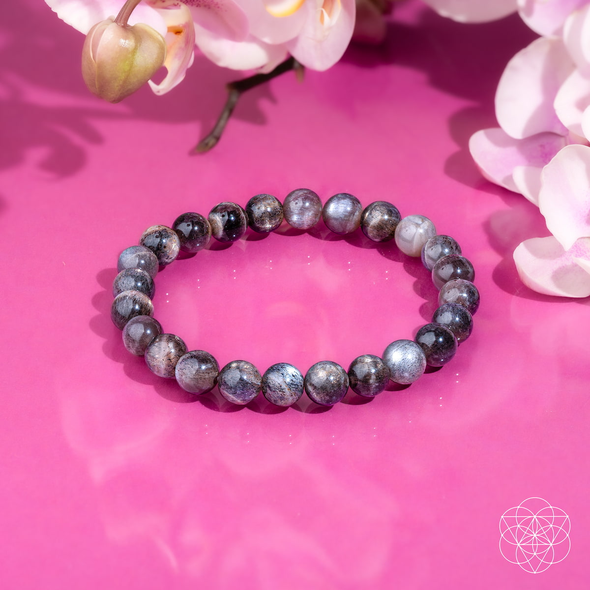 The Empath’s Cord Cutter – Black Moonstone Bracelet by Conscious Items, known for aiding new beginnings and energetic protection, is displayed on a pink surface with white and pink orchids. A geometric flower logo appears in the lower right corner.
