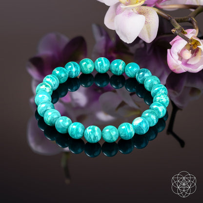 The Empire of Her Own – Premium Blue Amazonite Bracelet by Conscious Items, inspired by Amazon Warriors, is displayed on a glossy black surface with reflections beside pink and white orchids. A geometric white logo is in the lower right corner.