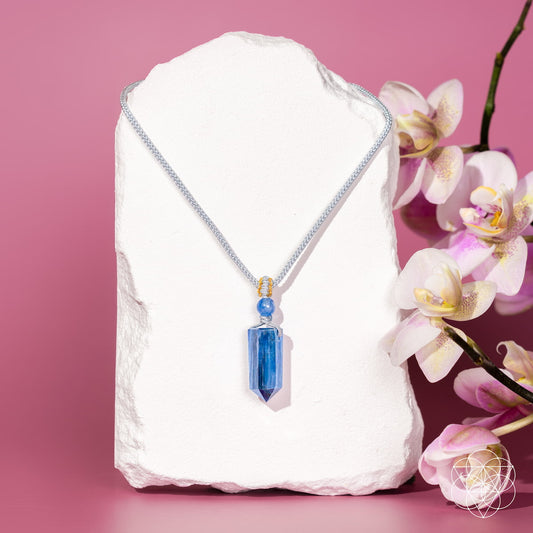 The Aura Shield - Rare Kyanite Pendant by Conscious Items features a blue kyanite stone on a silver chain, displayed with orchids on a pink background—ideal for energy resets and balancing the throat chakra.