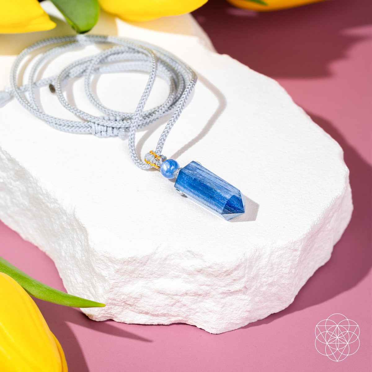 The Aura Shield - Rare Kyanite Pendant by Conscious Items rests on a white stone slab with yellow tulip petals, displayed on a pink background—designed to reset your throat chakra. A geometric white logo appears in the lower right corner.