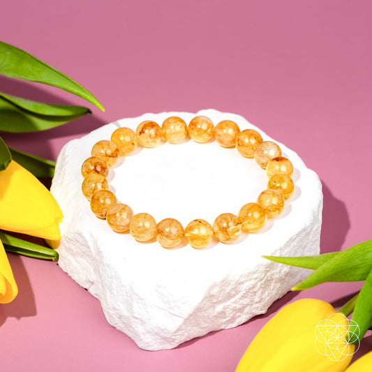 The Master Reset – Golden Healer Quartz Bracelet by Conscious Items is displayed on a white stone platform with yellow tulips and green leaves, set against a pink background—ideal for those seeking the Master Healer crystal.