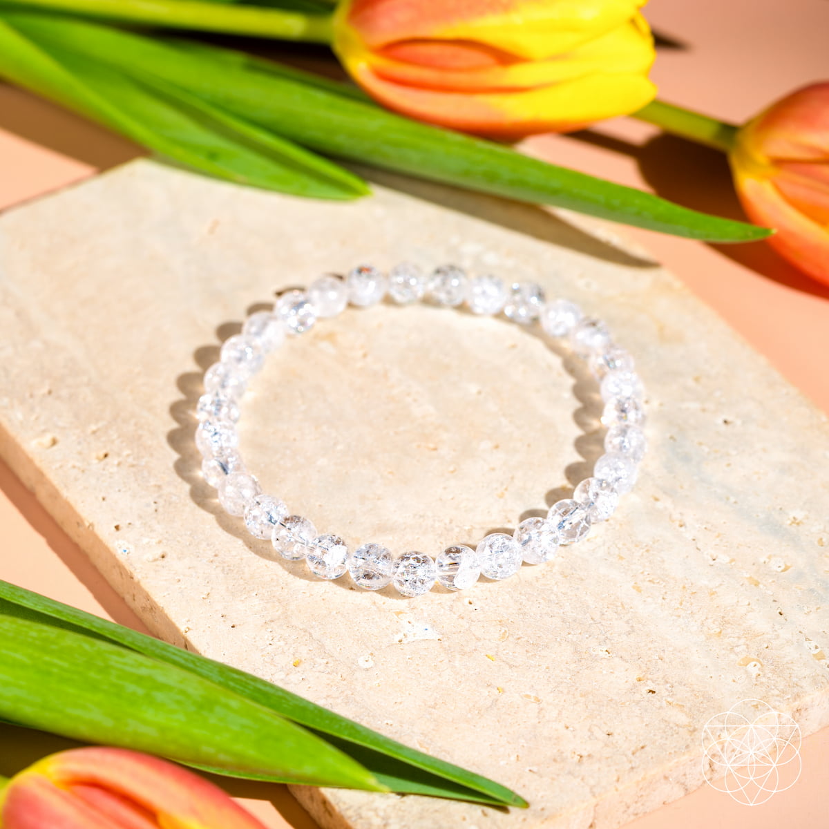 The Emotional Ice Bath – The Ice Quartz Bracelet | 6mm by Conscious Items, a reset stone for emotional clarity, is displayed on a beige stone slab with green leaves and orange-yellow tulips, sparkling in bright natural light.