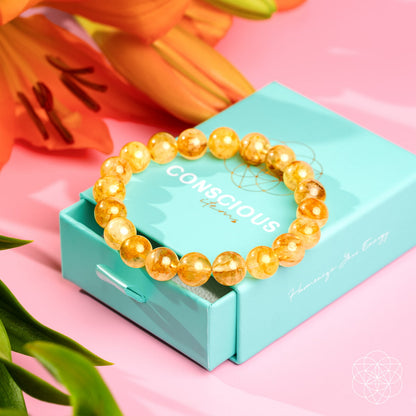 The Master Reset – Golden Healer Quartz Bracelet by Conscious Items lies on a turquoise jewelry box, with orange lilies and green leaves in the background, set against a pink surface.