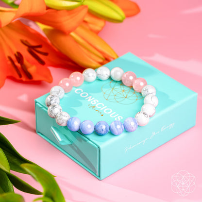 The 3AM Thoughts Off - Deep Sleep Trio Bracelet by Conscious Items, featuring soothing white, pink, and blue beads, is displayed on a turquoise Conscious jewelry box with orange lilies and green leaves on a pink background.