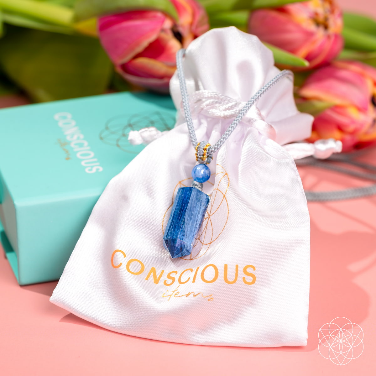 The Aura Shield - Rare Kyanite Pendant by Conscious Items, a silver chain necklace with a blue Kyanite stone, rests on a satin pouch. Pink tulips and a turquoise gift box create a perfect setting for promoting throat chakra balance and energy renewal.