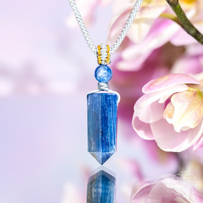 The Aura Shield - Rare Kyanite Pendant by Conscious Items, a silver necklace with blue kyanite and gold accents, is showcased among pink and white flowers—ideal for subtle energy resets and throat chakra support.