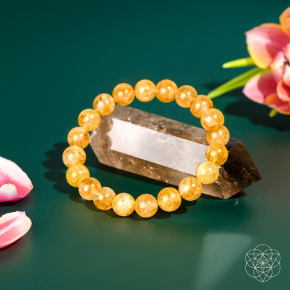 The Master Reset – Golden Healer Quartz Bracelet by Conscious Items rests on a polished crystal, surrounded by pink flowers on a green backdrop. A geometric white logo is visible in the corner.