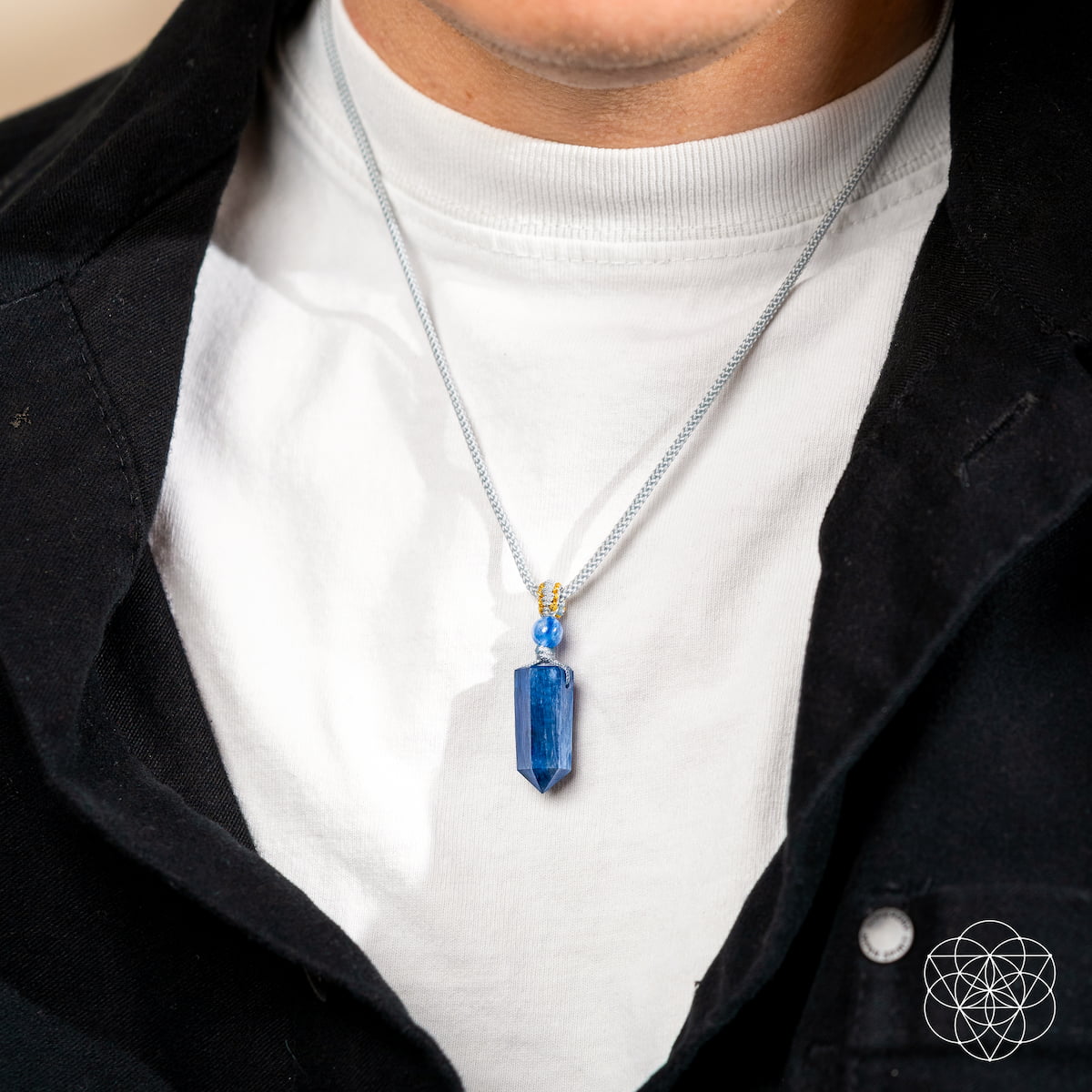 A person in a white crew-neck shirt and black jacket is shown close-up, wearing The Aura Shield - Rare Kyanite Pendant by Conscious Items—a silver-chained necklace ideal for supporting throat chakra balance and energy reset.