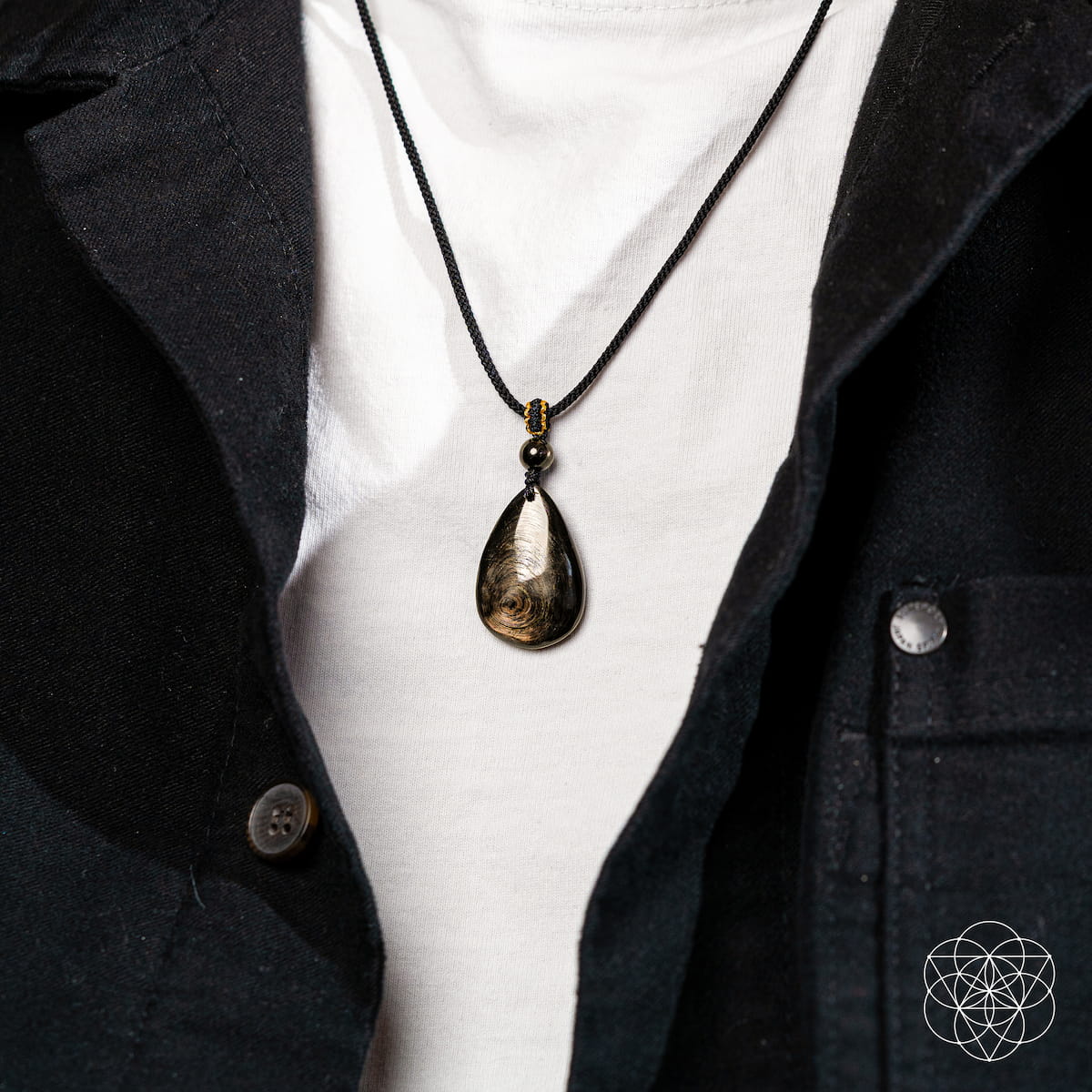 A person in a white shirt and black jacket, shown from the shoulders down, wears The Hypersthene Millionaire Manifestation Pendant by Conscious Items on a black cord. A geometric logo appears in the bottom right corner.