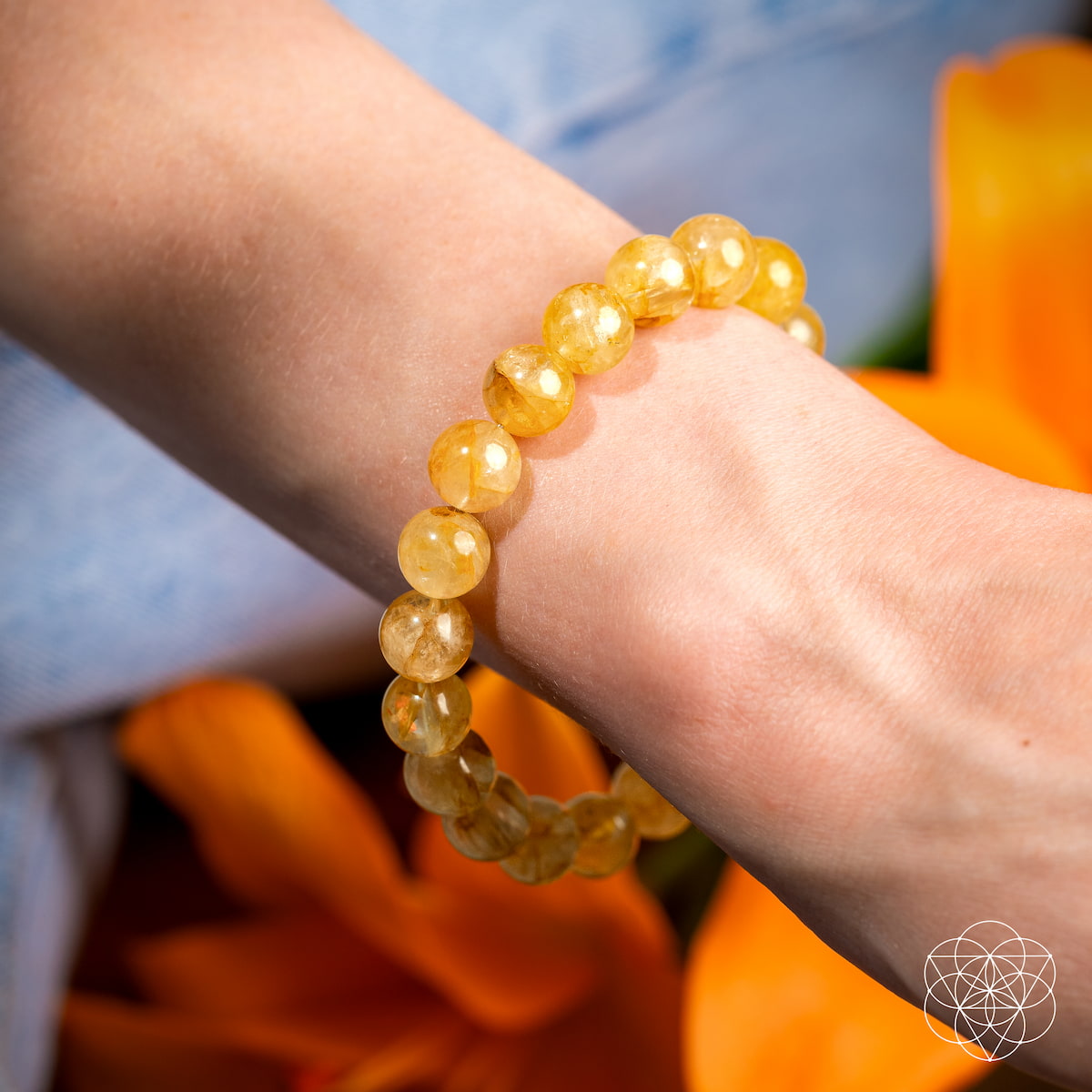 A wrist wears The Master Reset – Golden Healer Quartz Bracelet by Conscious Items, featuring yellow beads. Orange flowers and a blue garment are blurred in the background, with a white geometric logo at the bottom right.