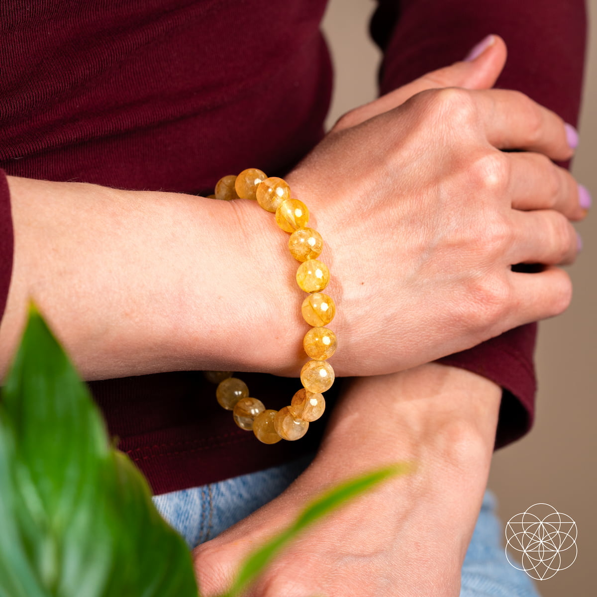 A person wears The Master Reset – Golden Healer Quartz Bracelet by Conscious Items—a vibrant yellow beaded accessory—paired with a maroon top, light blue jeans, green leaves in the foreground, and a geometric white logo in the corner.