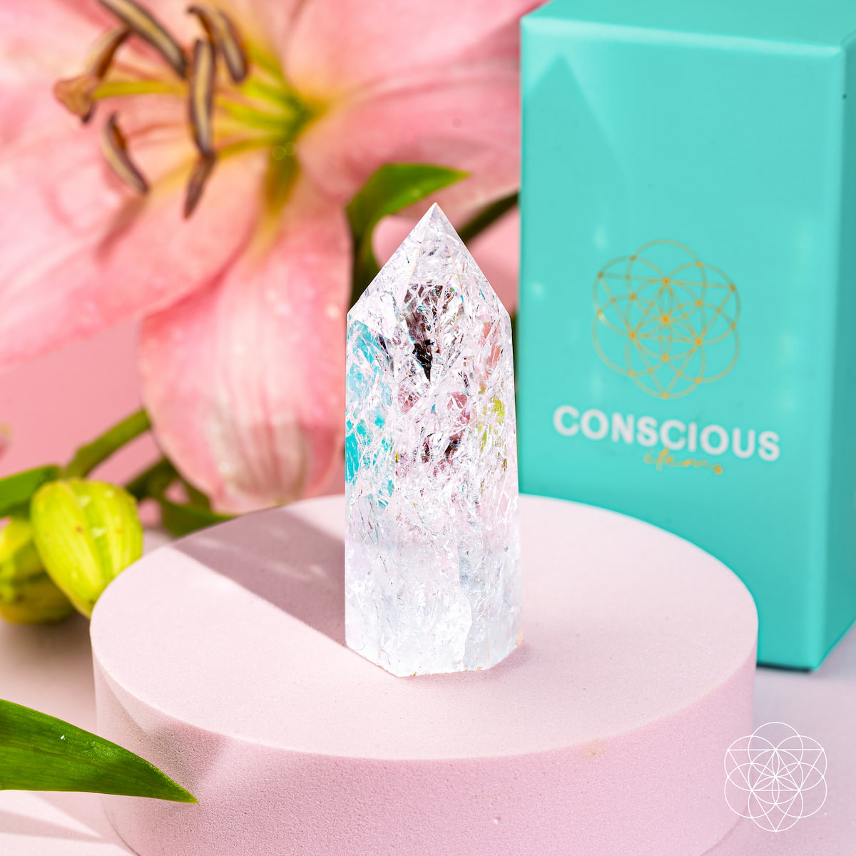 The Limitless Manifestation - Ice Quartz Point by Conscious Items stands on a round pink platform with a pink flower and a turquoise Conscious box in the background. Vivid reflections within the crystal amplify its manifestation energy.